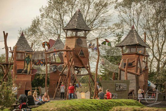 Playground with wooden castle towers, rope bridges, and children playing outdoors.