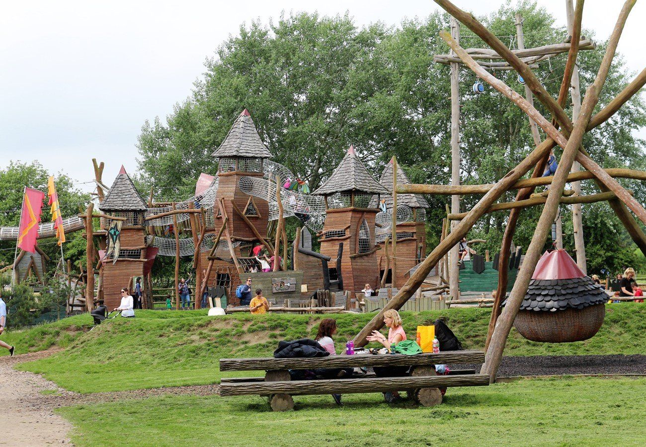 Playground with wooden castle, picnic area, and swings. Children and adults in green space on a cloudy day.