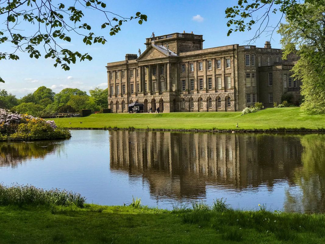 Large stone mansion reflected in a pond, set in a grassy landscape with trees.