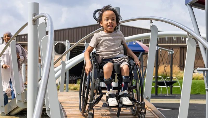 Boy in wheelchair on playground bridge with adult in background.