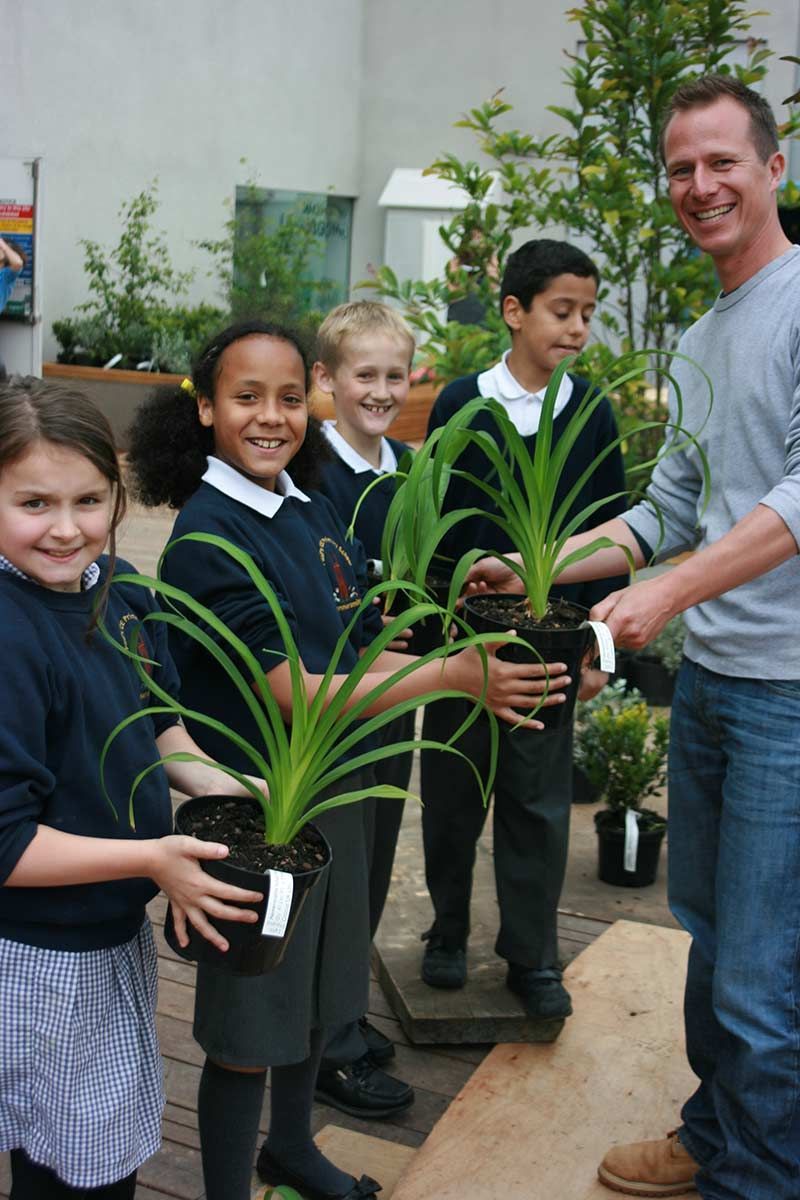 Schoolchildren holding potted plants with a smiling man, outdoors.