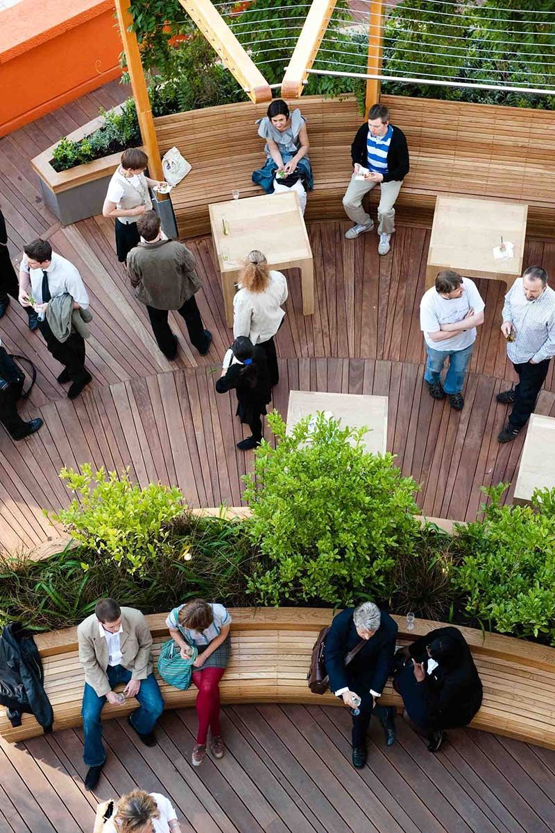 People socialize and relax on a wood deck with benches and tables. Sunny day.