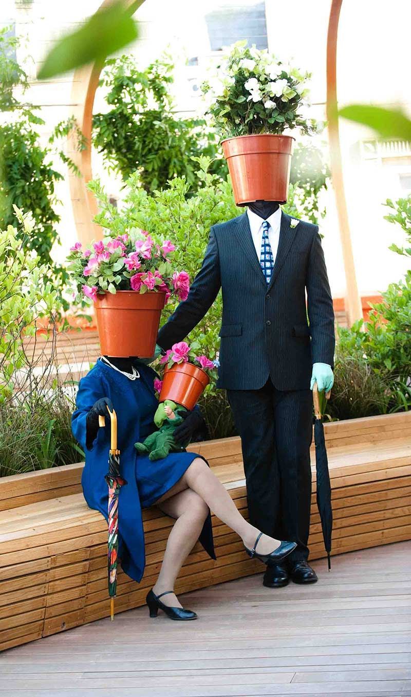 Two figures, heads replaced by flower pots, posing on a wooden bench. One in a suit, one in a dress. Garden setting.
