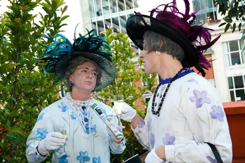 Two people in drag as elderly women, wearing large hats, dresses with floral print, outdoors, talking.