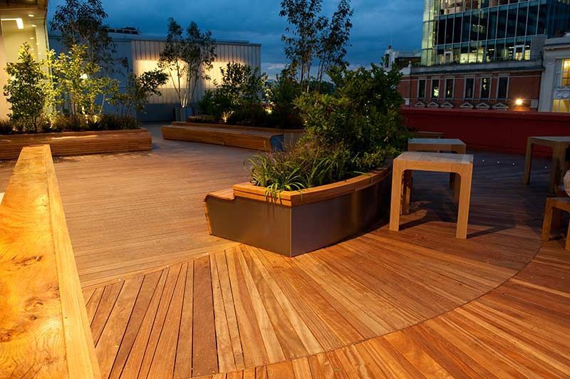Rooftop patio with wooden deck, planters, and city lights at dusk. Empty stools sit near the plants.