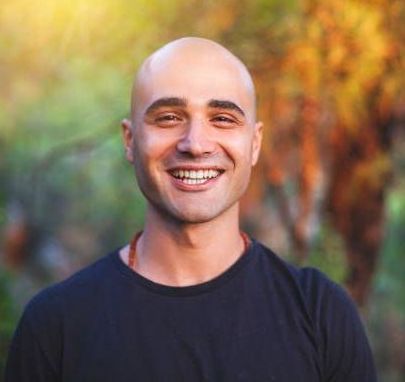 Bald man smiles at the camera, wearing a black shirt. Blurred background of a sunlit forest.