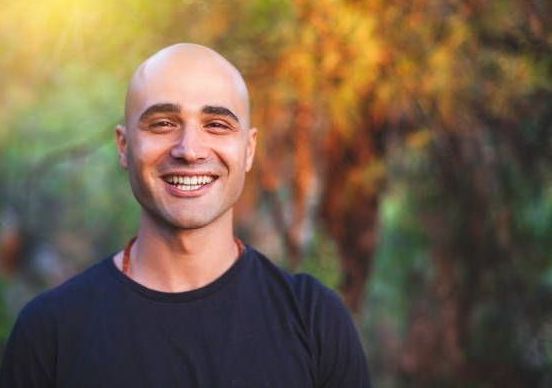 Bald man smiles widely, outdoors; blurred foliage in background.