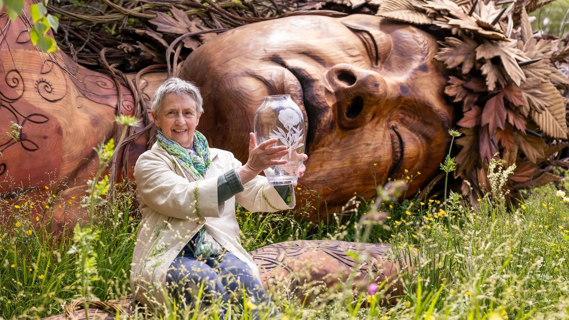 Sarah Eberle - holding a clear glass trophy in front of a large carved wooden face surrounded by greenery