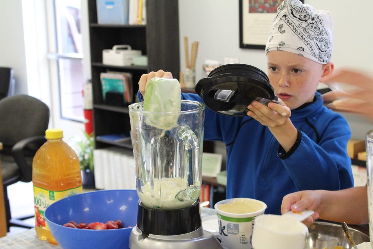 Child in a bandana pours ingredients into a blender on a kitchen counter with bowls and drinks nearby
