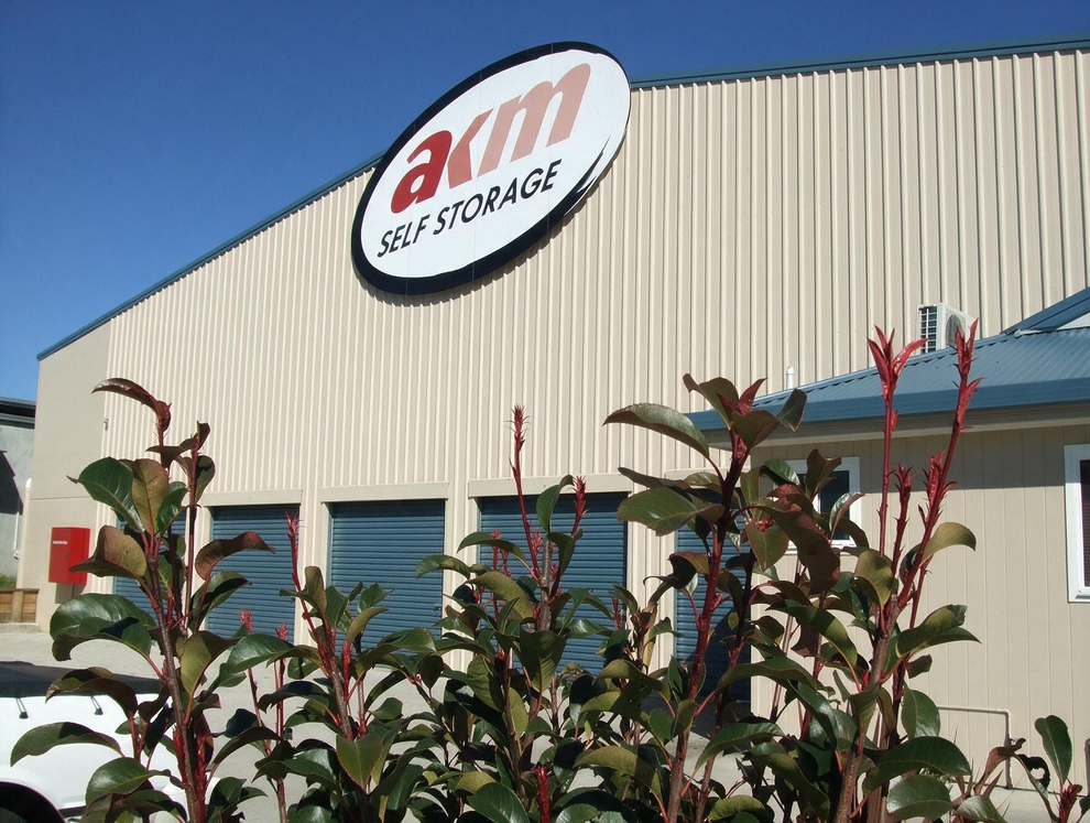 Industrial building with an oval AIM sign above blue garage doors, partly obscured by red-flowered shrubs.