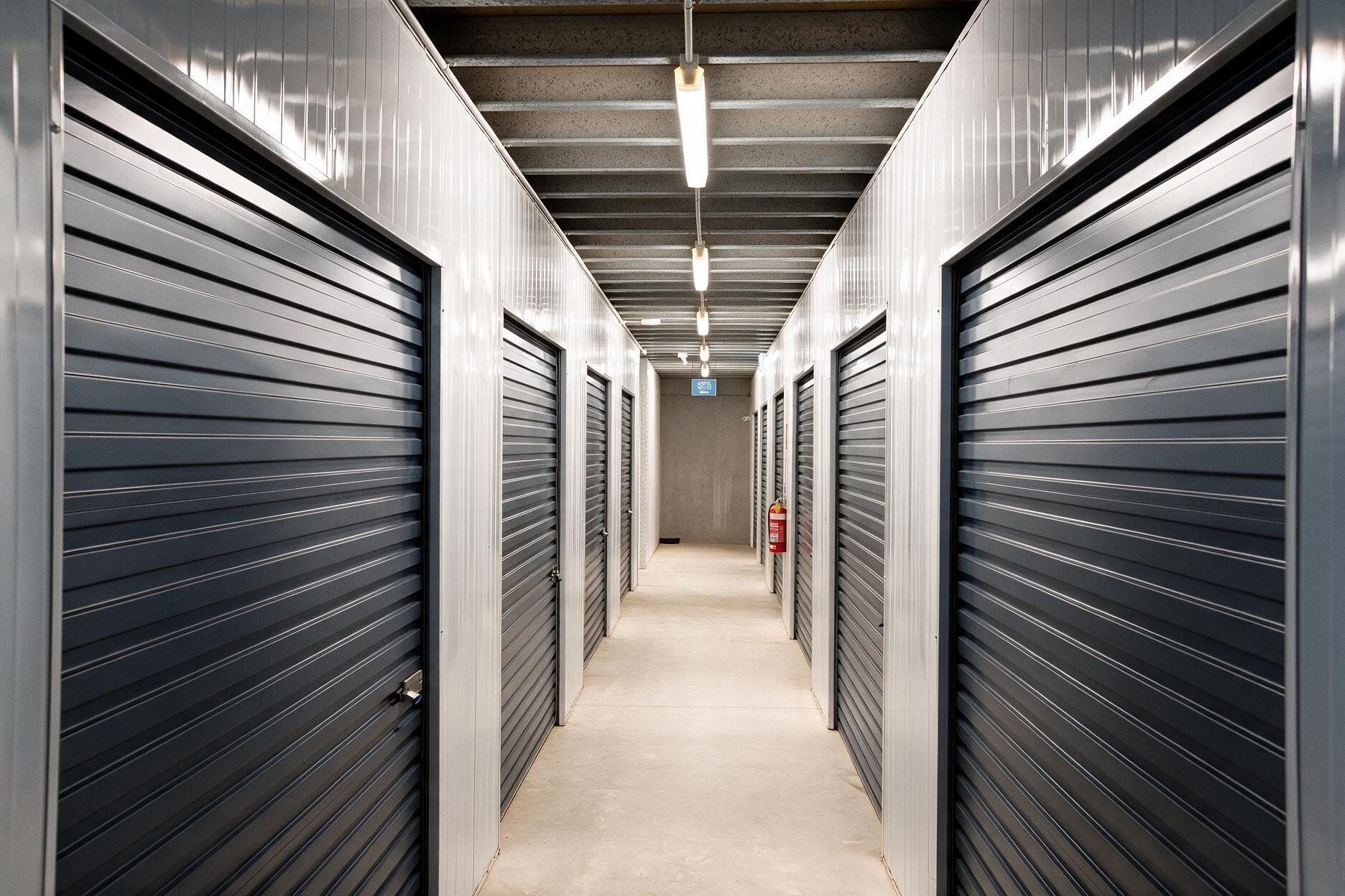 A row of gray storage unit doors in a narrow indoor hallway with bright overhead lights.