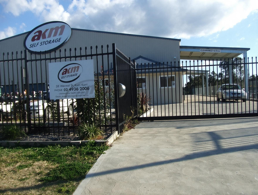 Car dealership entrance with black metal gate, white building, and parked cars under a covered lot.