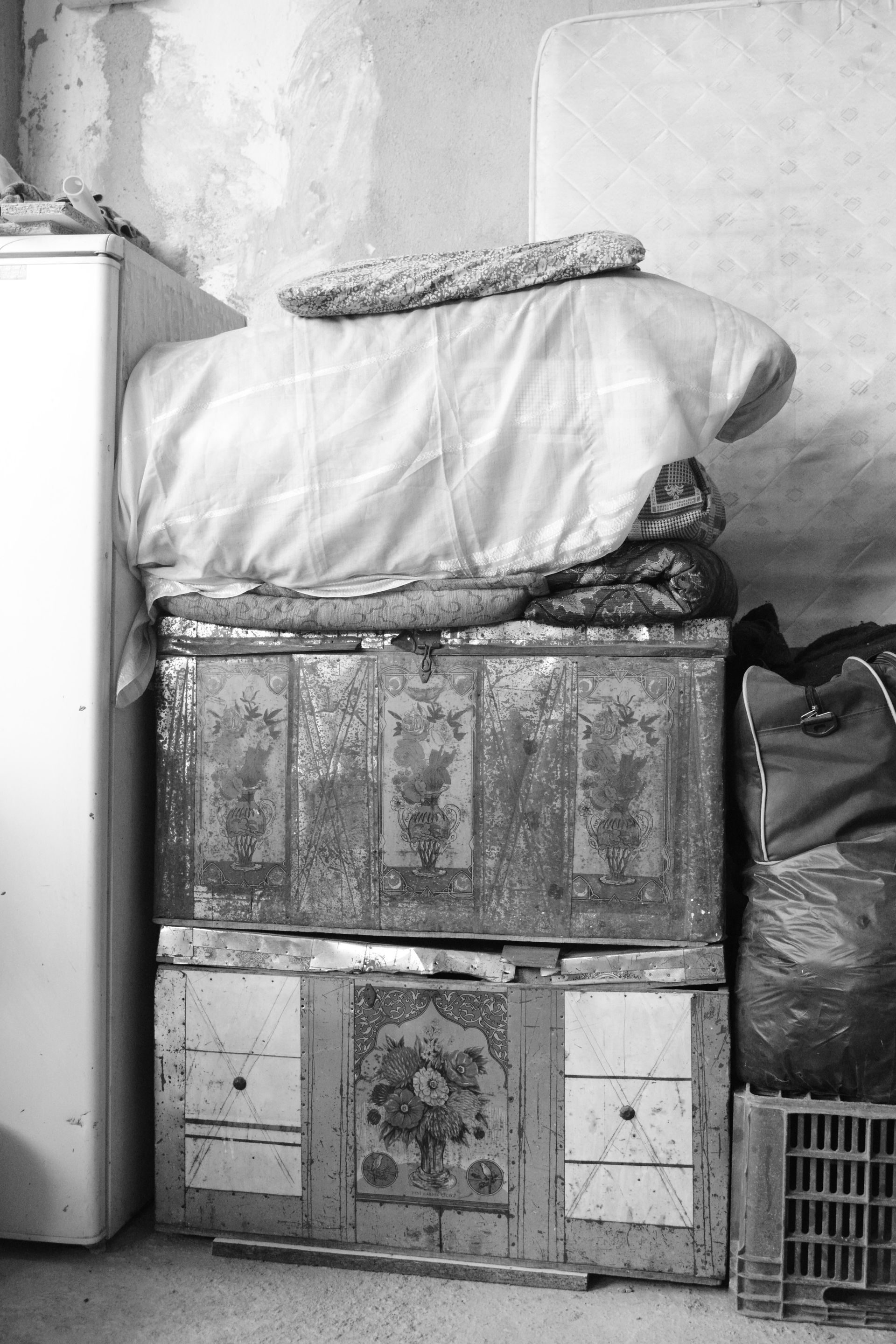 Stacked bedding and crates beside a wall, with a refrigerator and plastic bag in a cramped room.