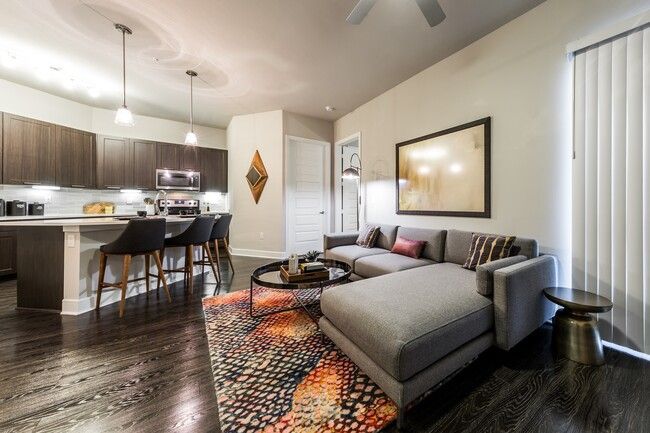 Open-concept living room with gray sectional, colorful rug, and kitchen bar stools beyond.