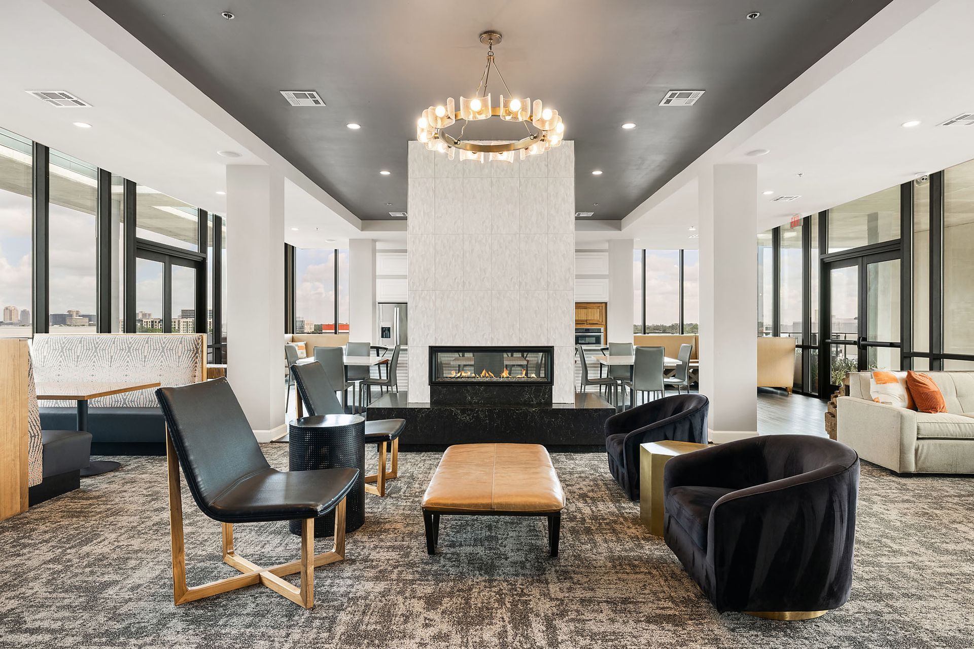 Modern living room with black chairs, ottoman, fireplace, and chandelier, framed by large windows.