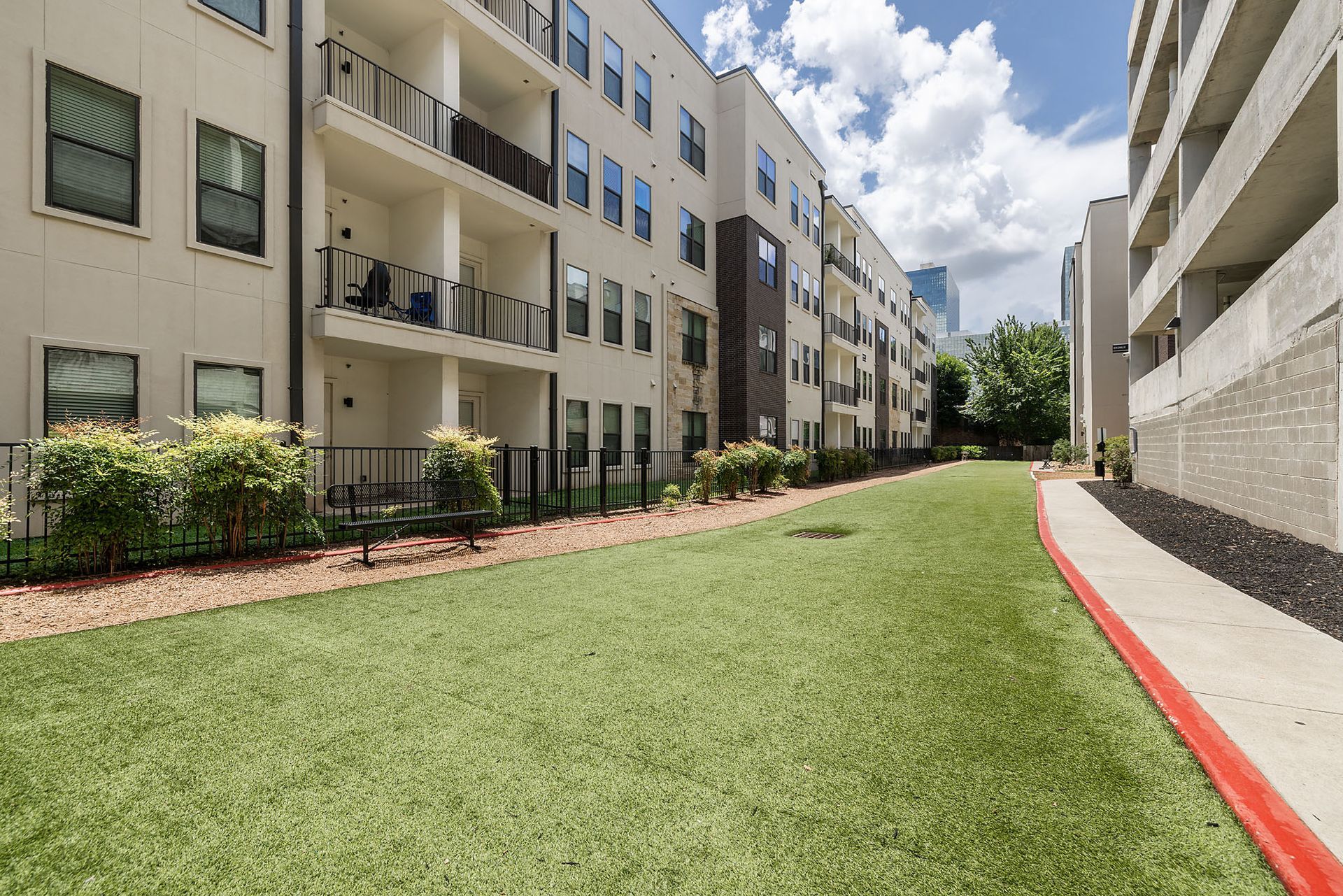 Apartment courtyard with green lawn between beige buildings and a red curb under a cloudy sky