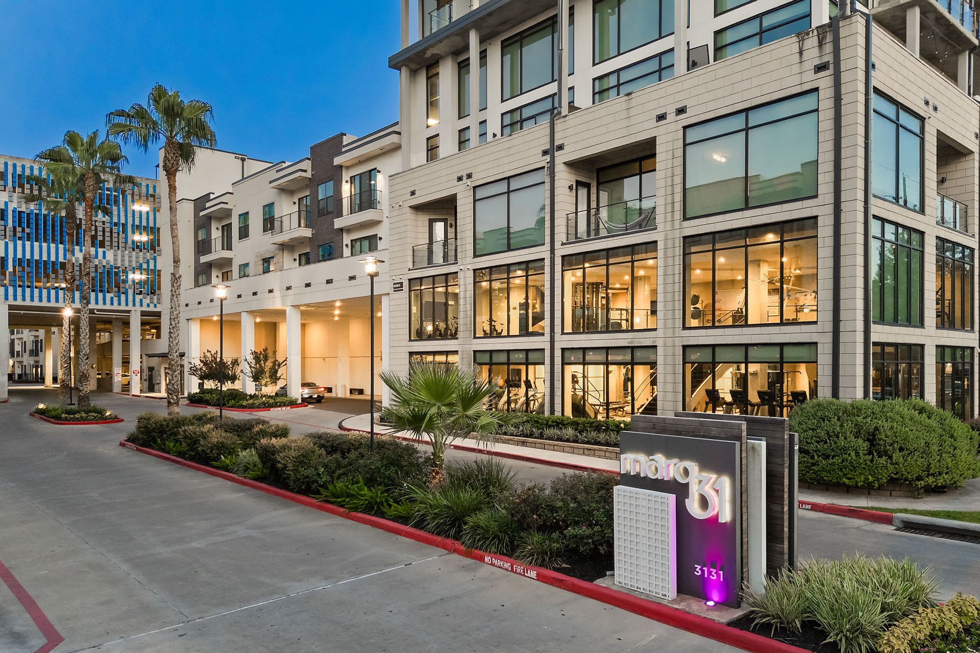 Modern apartment building with lit windows, landscaped entrance, and a street at dusk