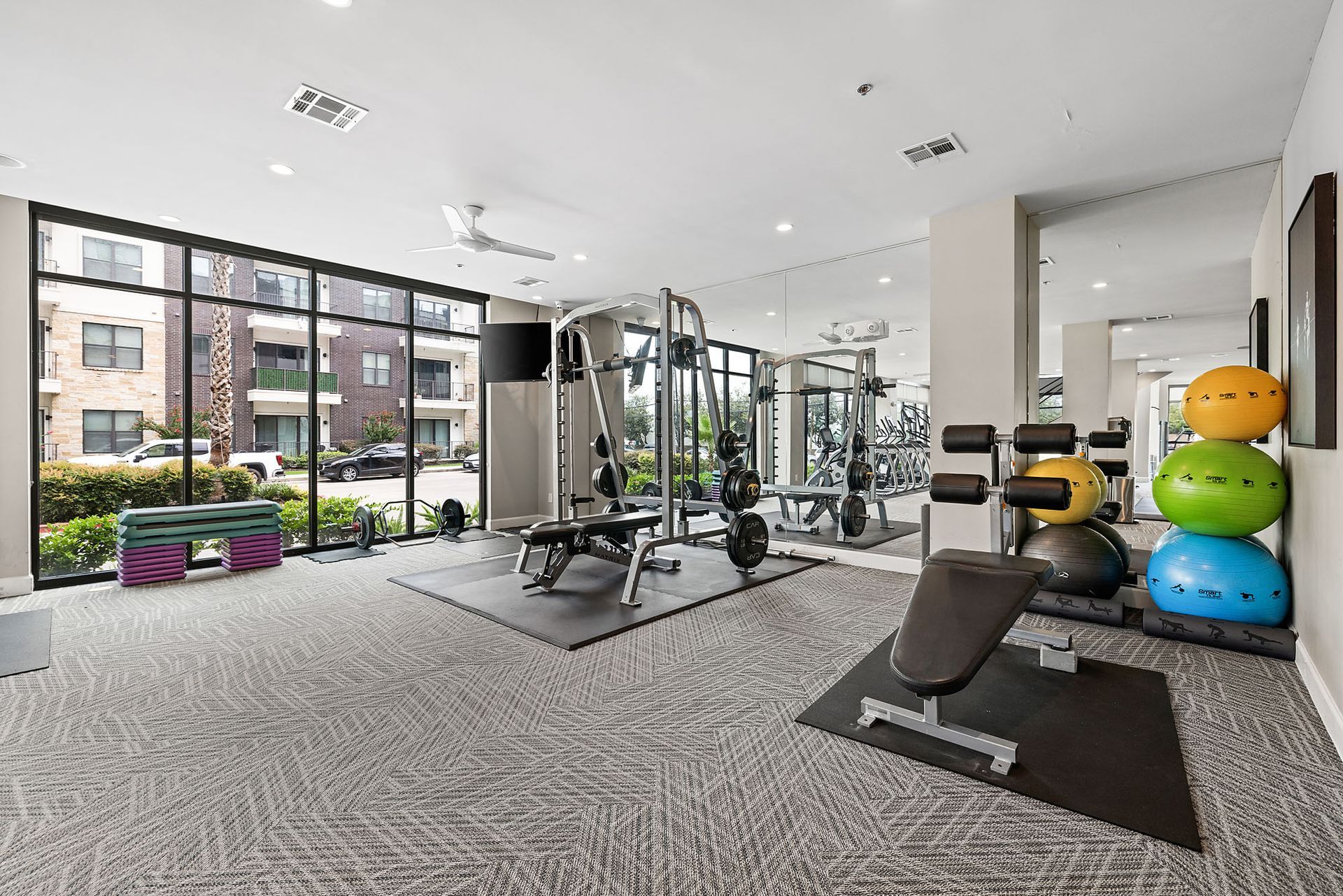 Modern gym with weights, benches, cable machine, and large windows overlooking apartments