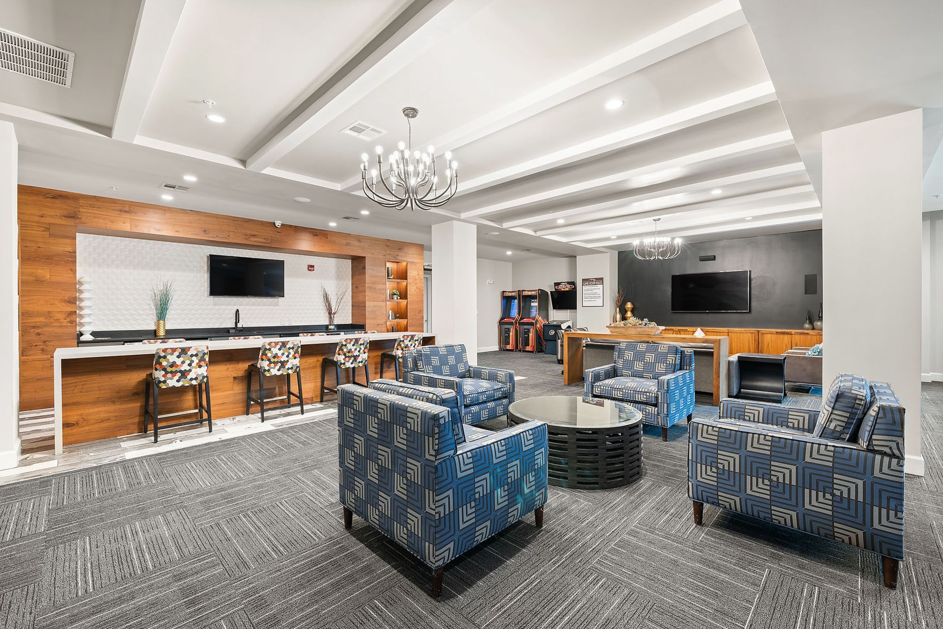 Modern lounge with blue patterned chairs, bar stools, TV screens, and a chandelier on a carpeted floor.