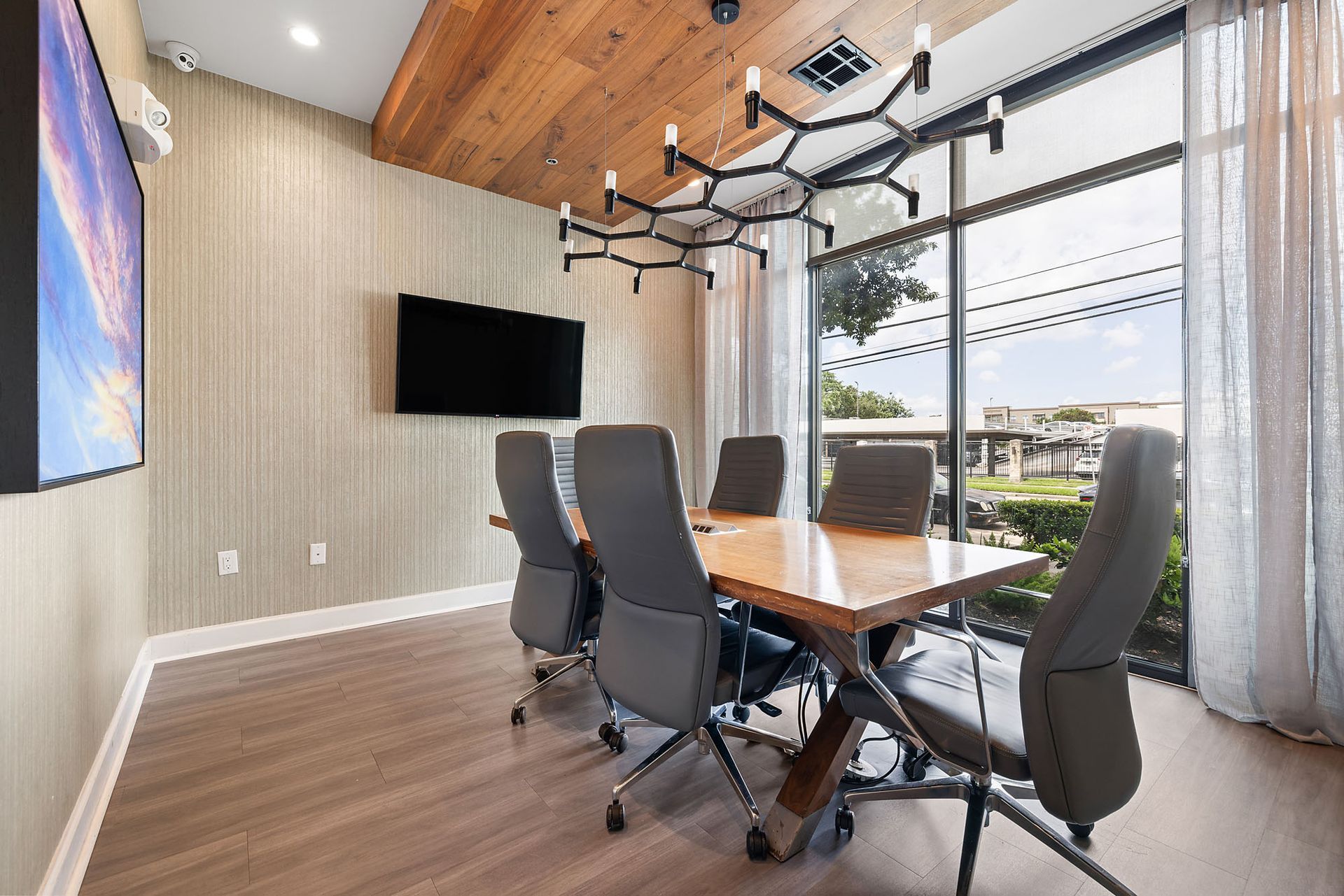 Modern meeting room with wood table, gray chairs, wall-mounted TV, and large windows overlooking a patio.