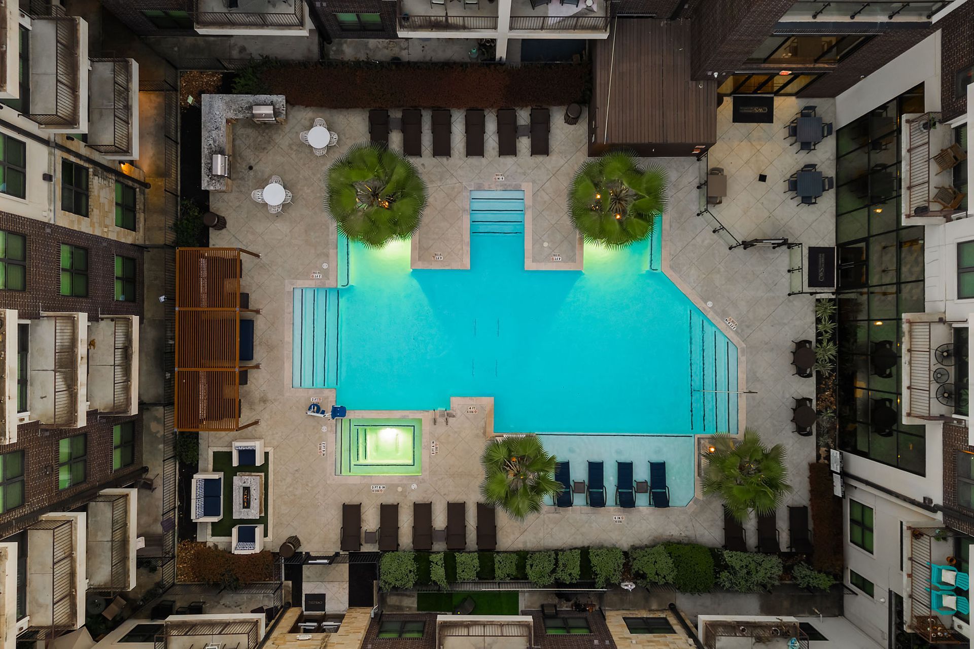 Aerial view of a turquoise swimming pool in a courtyard, surrounded by lounge chairs and trees.