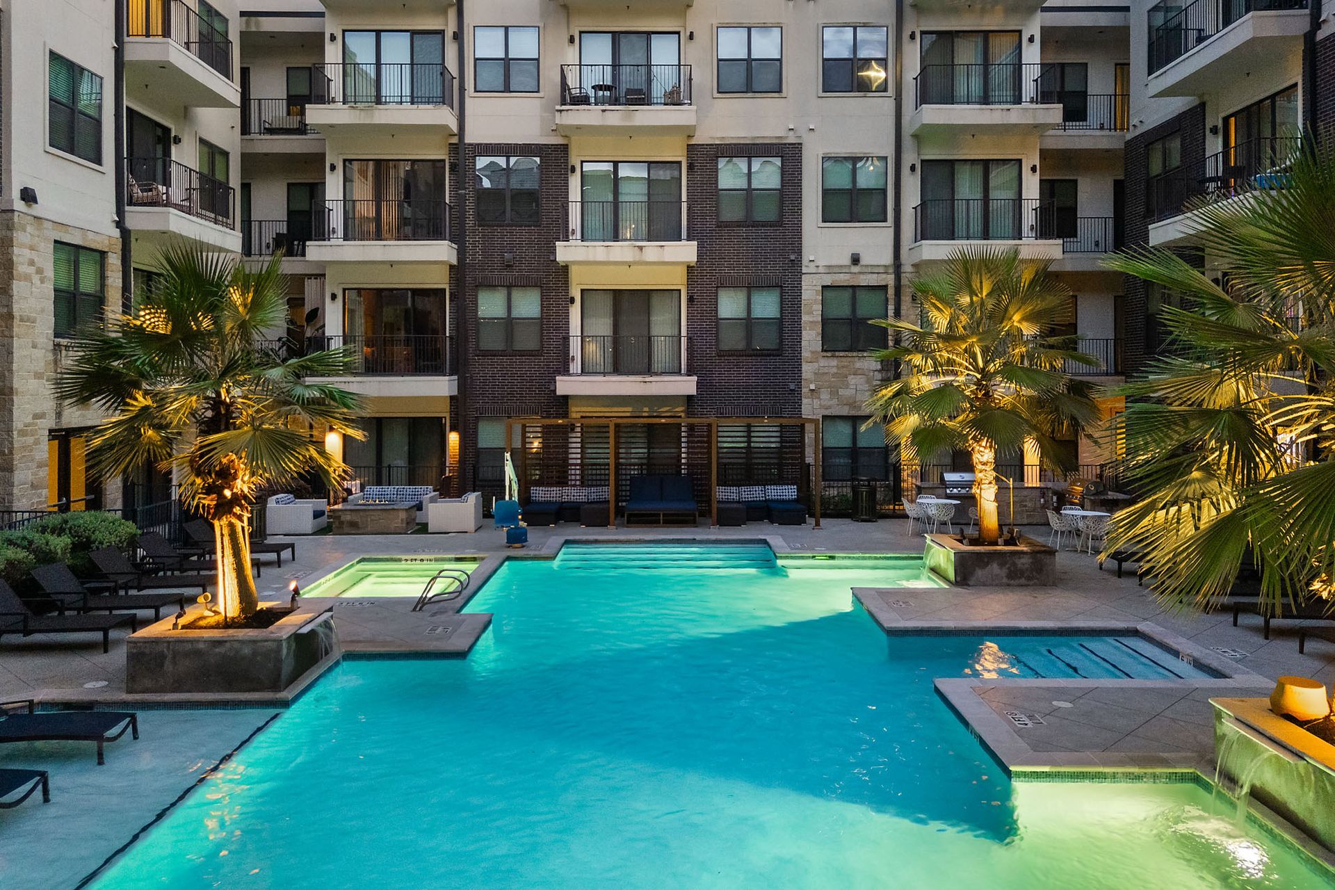 Courtyard apartment pool with turquoise water, lounge chairs, and illuminated palm trees at dusk