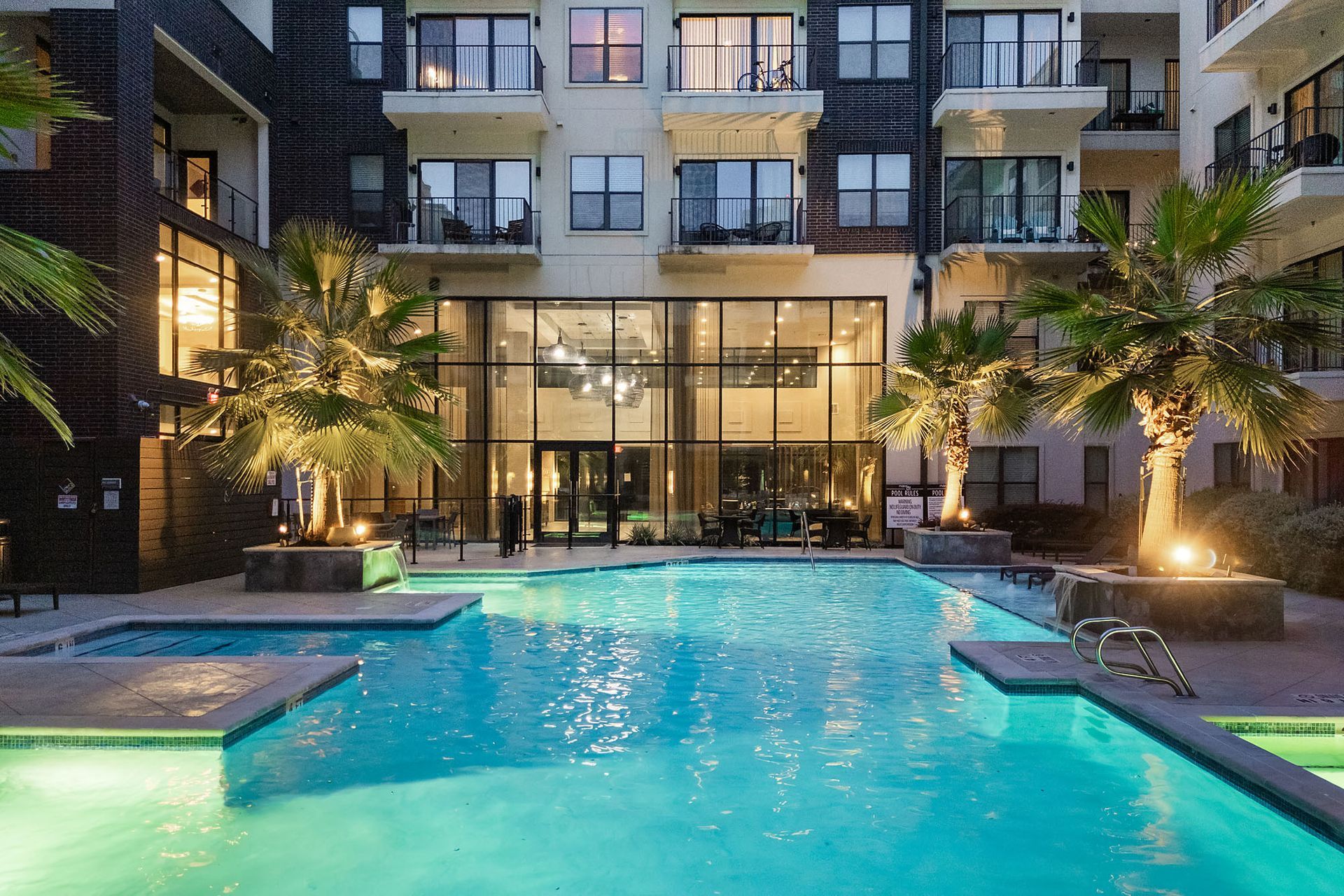Apartment courtyard with turquoise pool, palm trees, and lit lounge areas at dusk