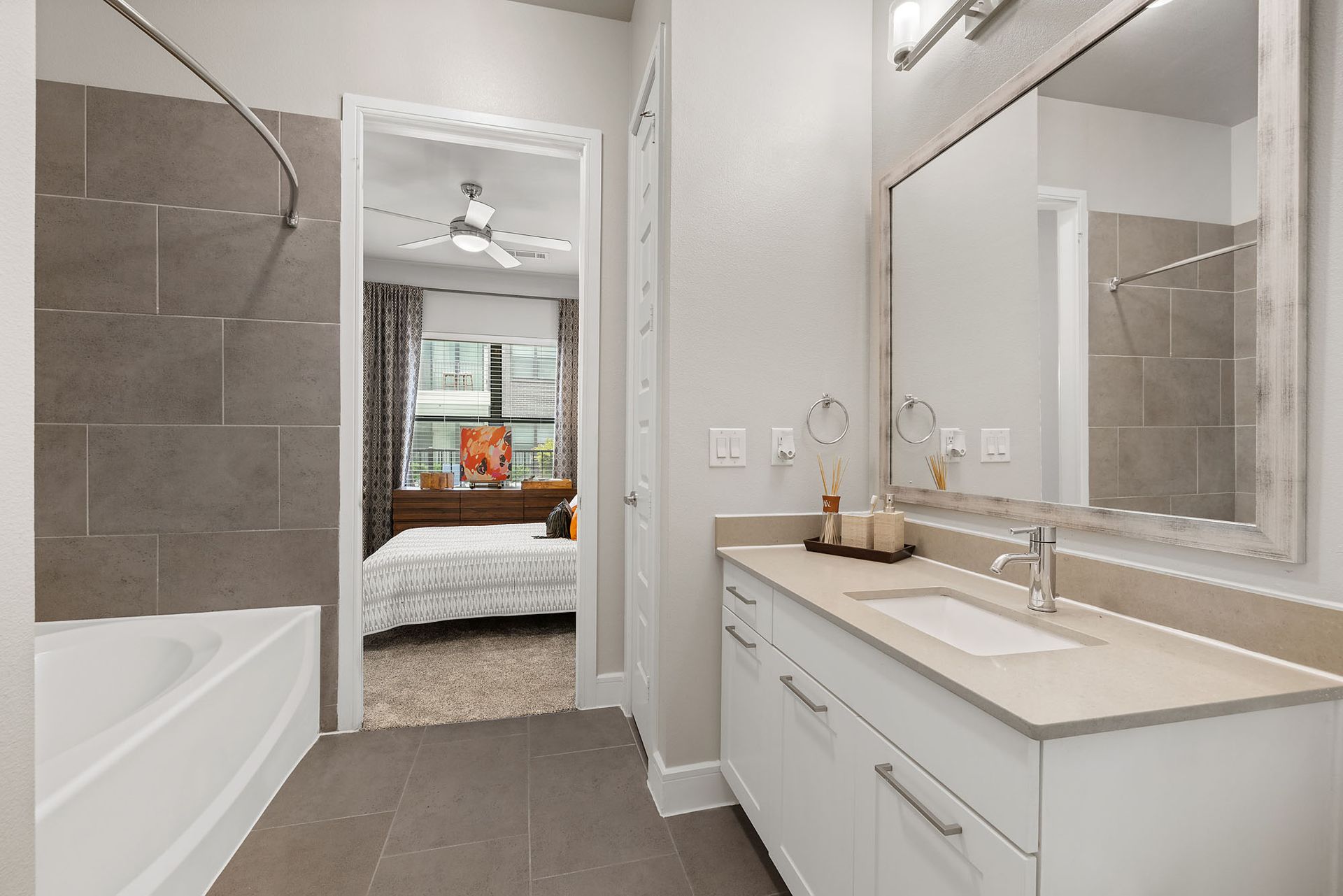 Modern bathroom with white vanity, large mirror, gray tile, and a view into a bedroom through the doorway