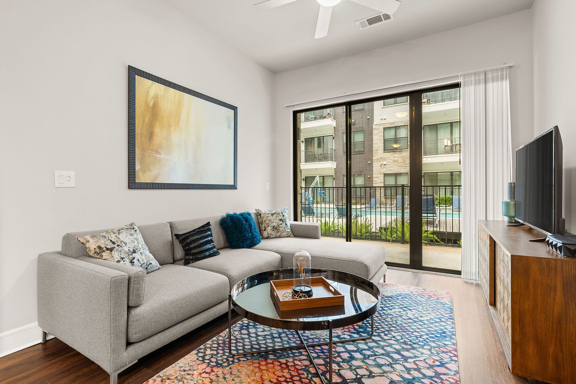 Modern living room with gray sectional, round coffee table, colorful rug, TV, and balcony doors.
