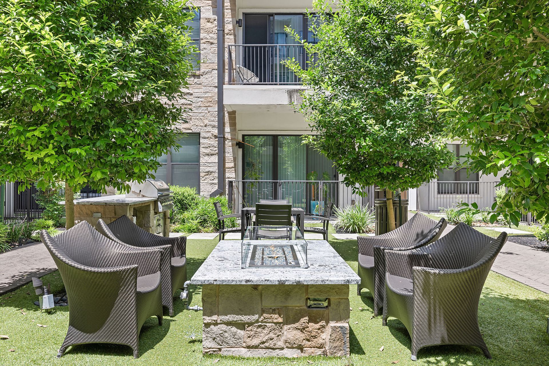 Courtyard patio with wicker chairs around a fire pit, surrounded by green trees and apartment balconies