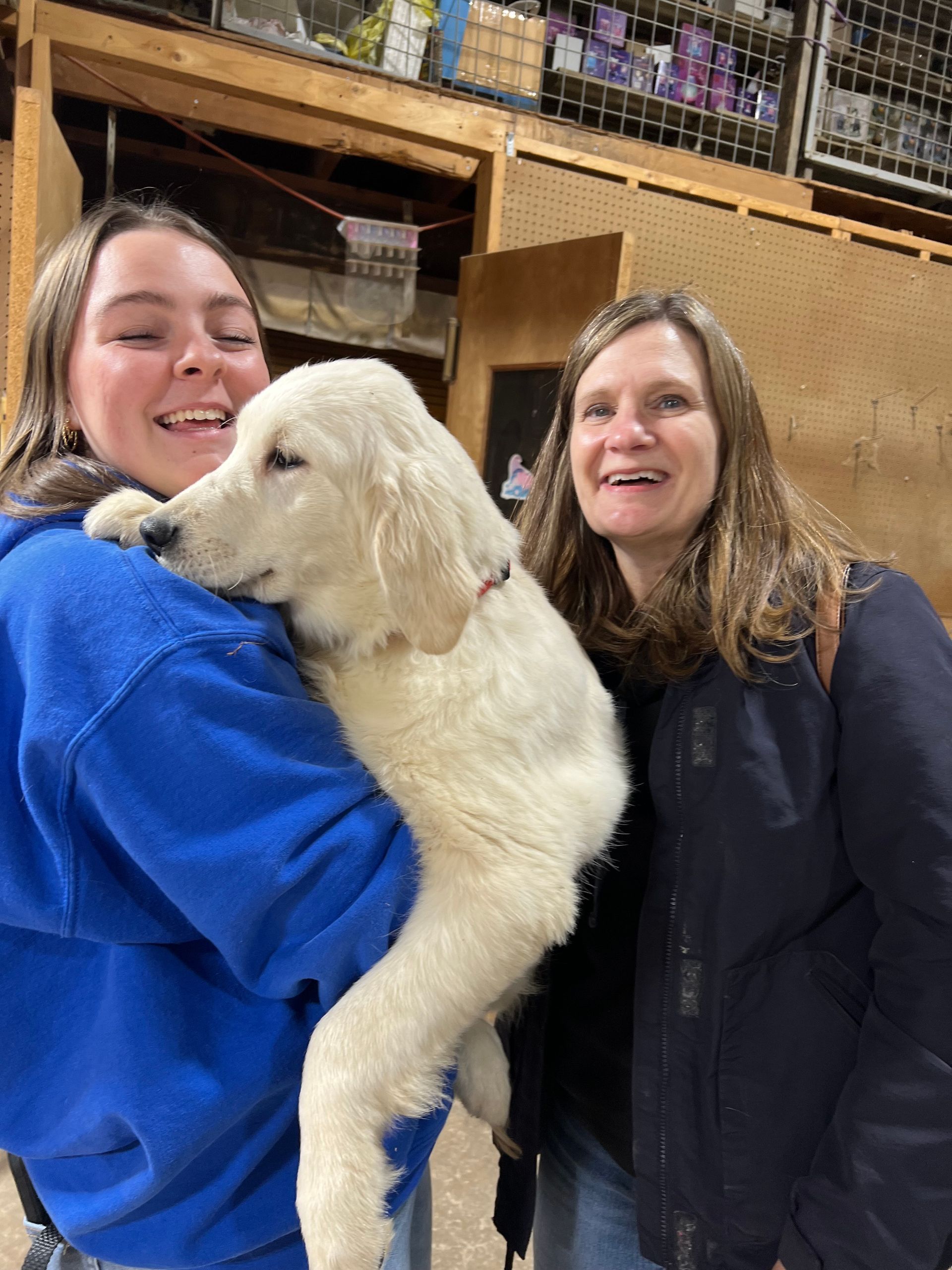 Two people smiling while holding a fluffy white puppy indoors