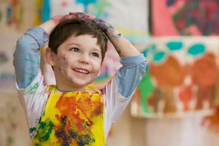 A young boy wearing a yellow apron is covered in paint.
