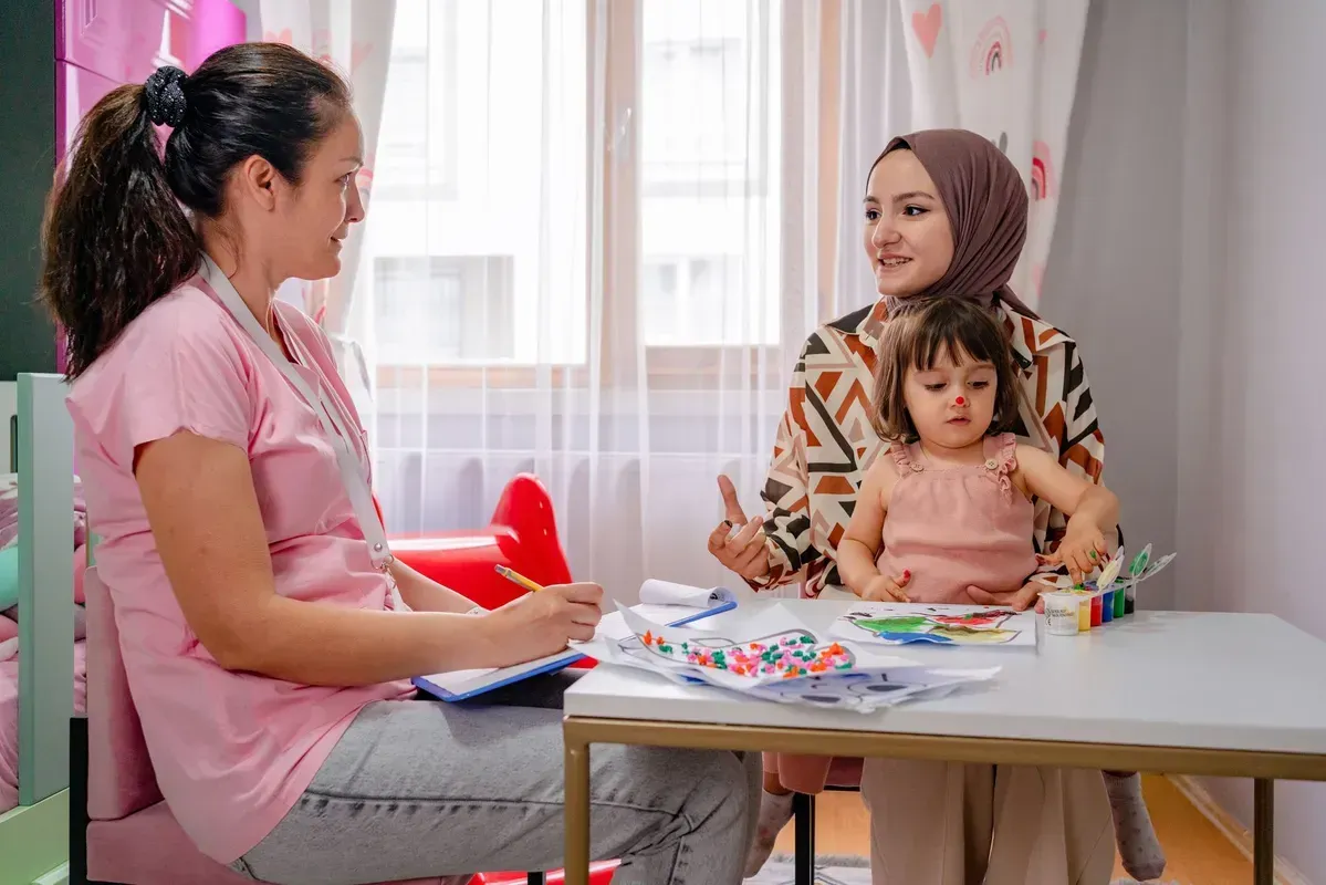 A woman is sitting at a table with a little girl and talking to her.