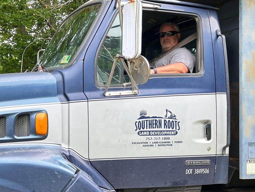 Man in sunglasses driving a blue-and-white Southern Roots truck, seen through the open window.