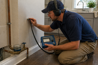 Technician kneels by a wall, using a water leak detector near exposed pipes.