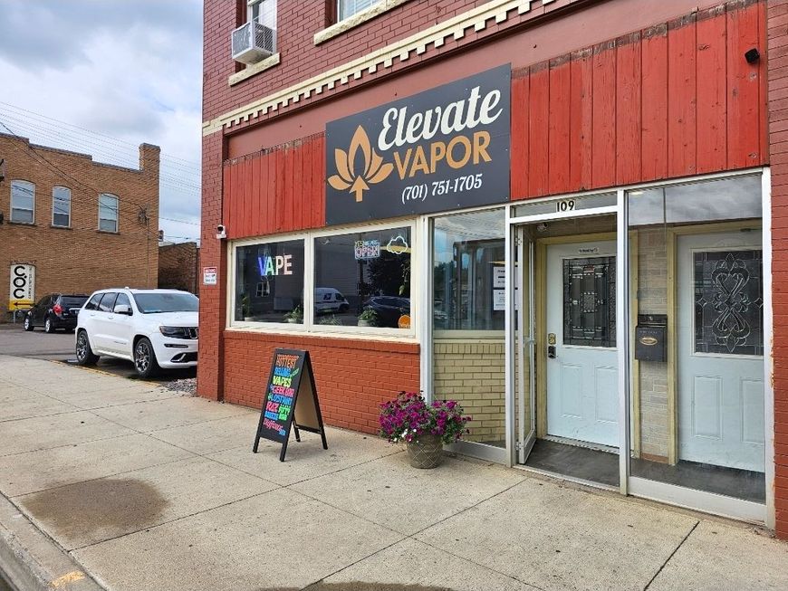 An Elevate Vapor shop exterior with a red brick facade, storefront windows, and a sidewalk sign in an urban setting.
