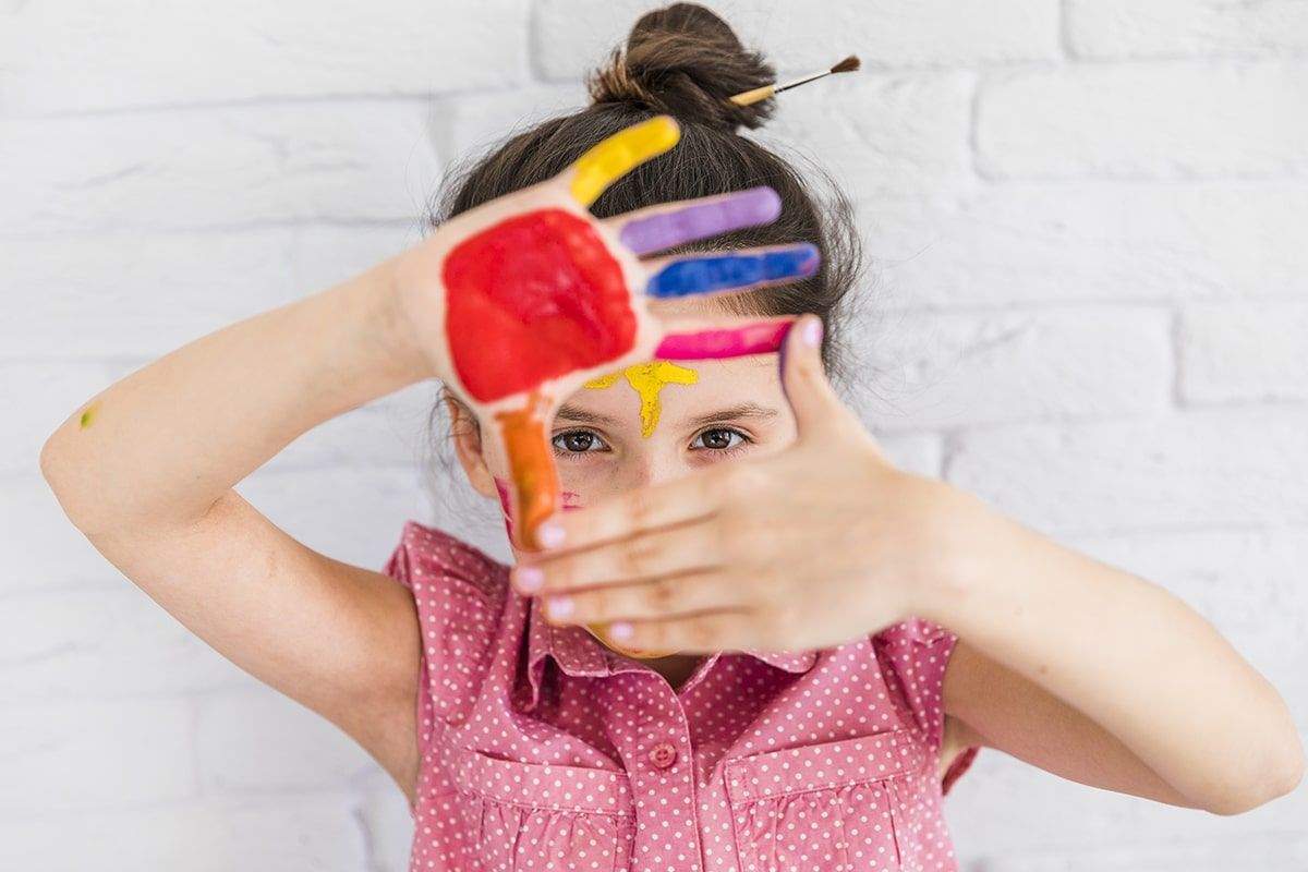 Uma menina está cobrindo o rosto com as mãos pintadas em cores diferentes.