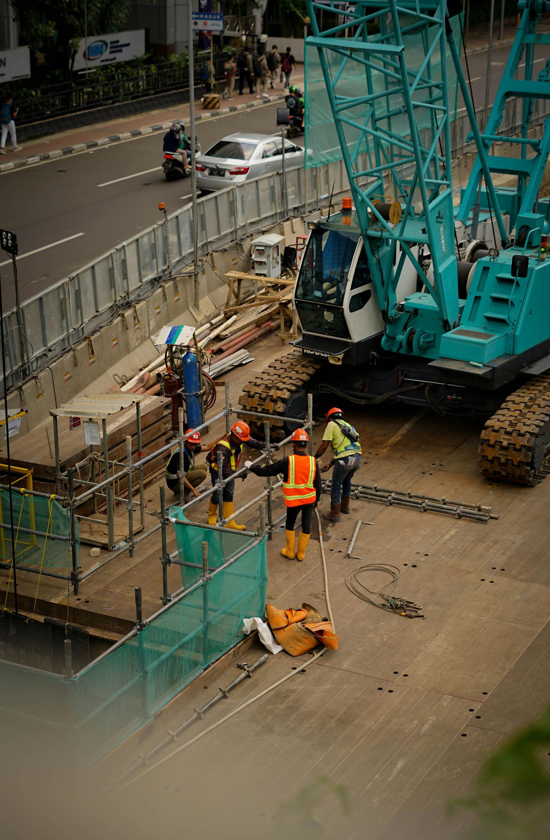 Construction site with workers, crane, and barrier on a city street. Workers wearing hard hats and vests.