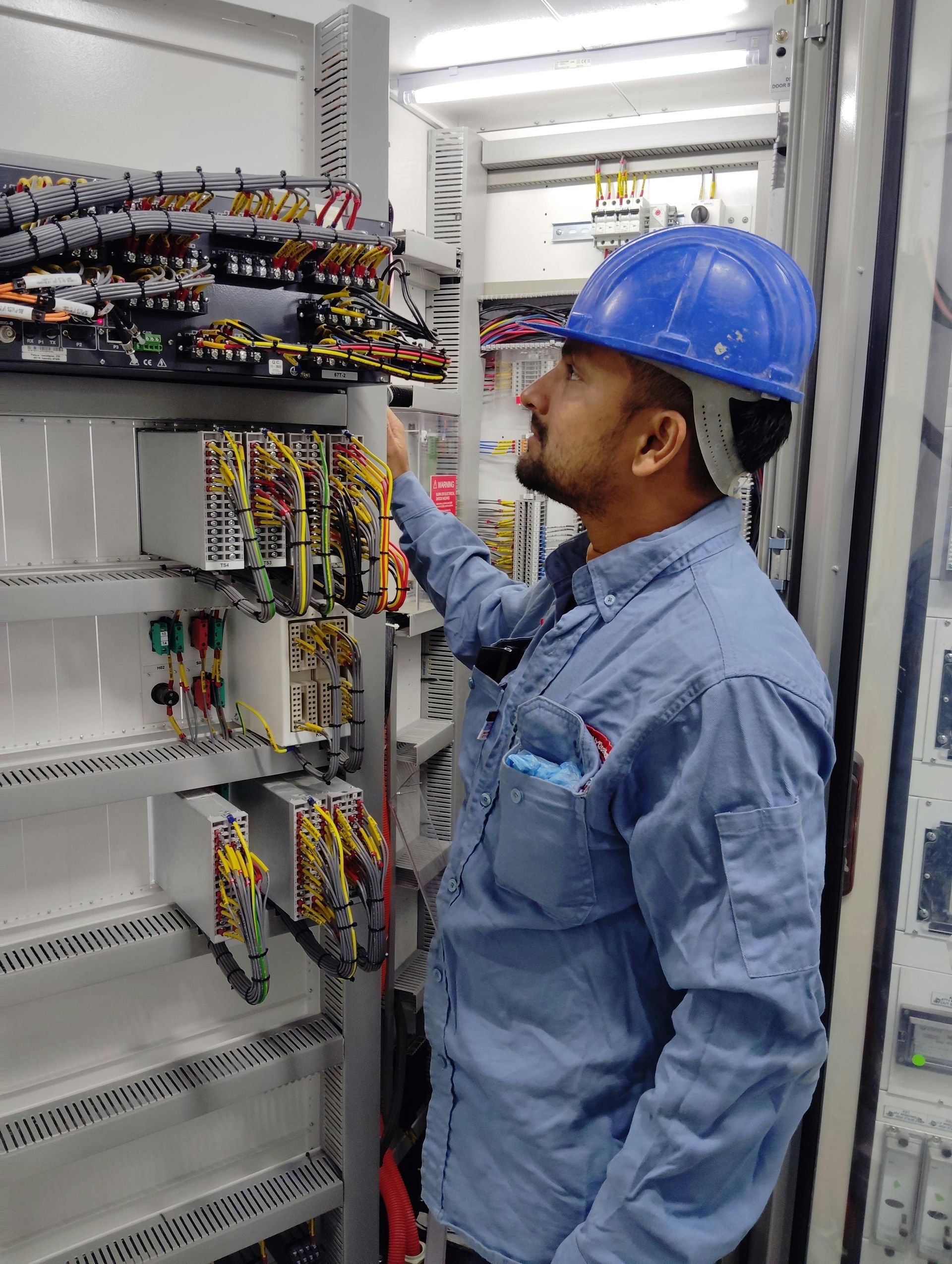 Man in blue hard hat examines electrical panel's wiring in a utility room.