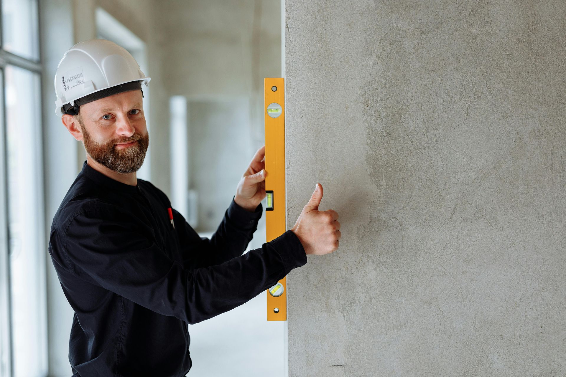 Construction worker with white hard hat using a level on a wall, giving a thumbs-up.