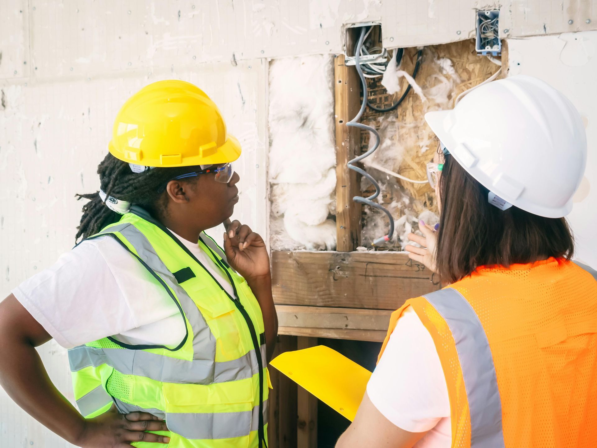 Two people in hard hats and safety vests inspect electrical wiring in a wall.