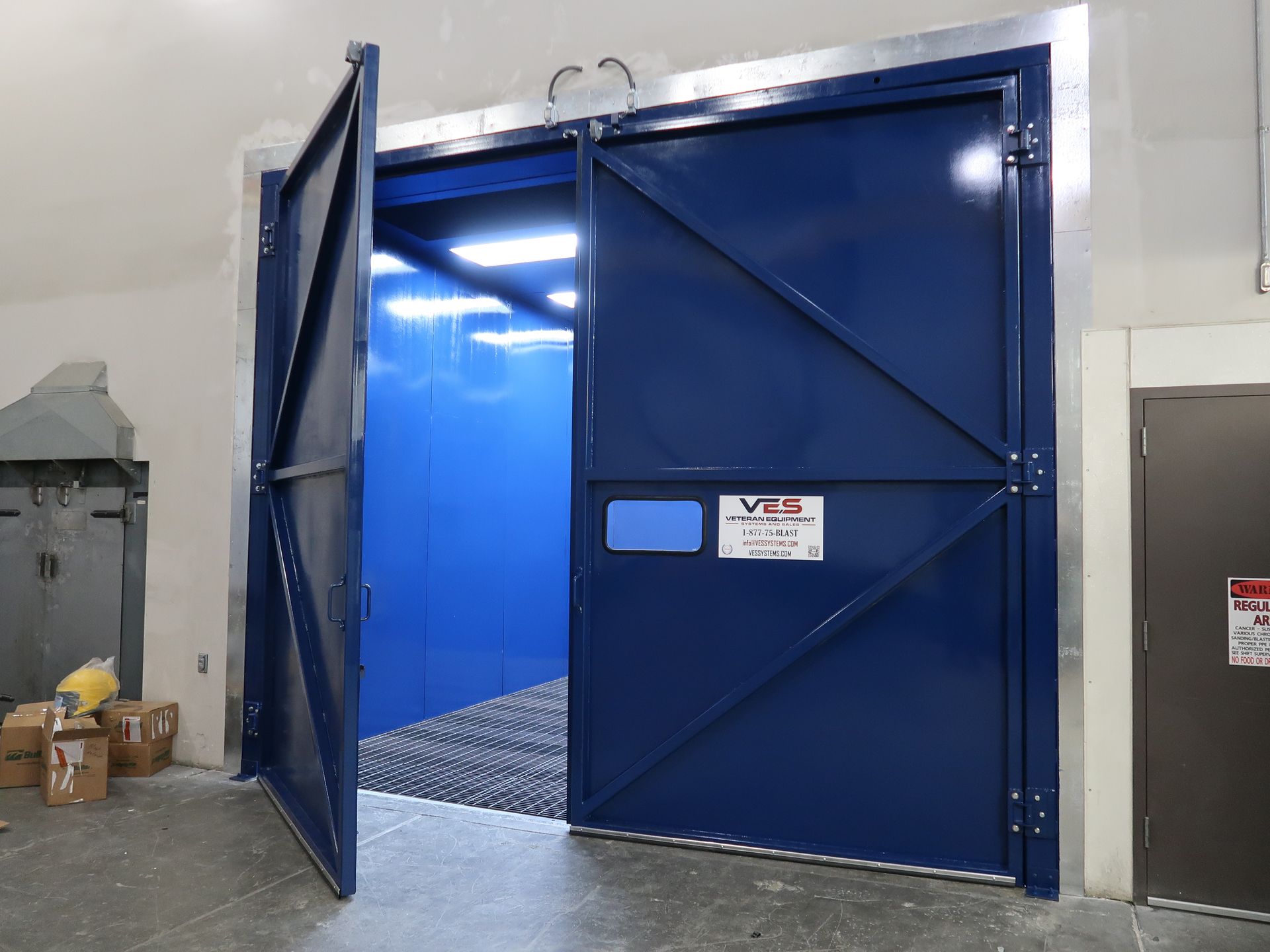 Blue VES blast enclosure with one door open showing the interior with blue walls and grated floors.