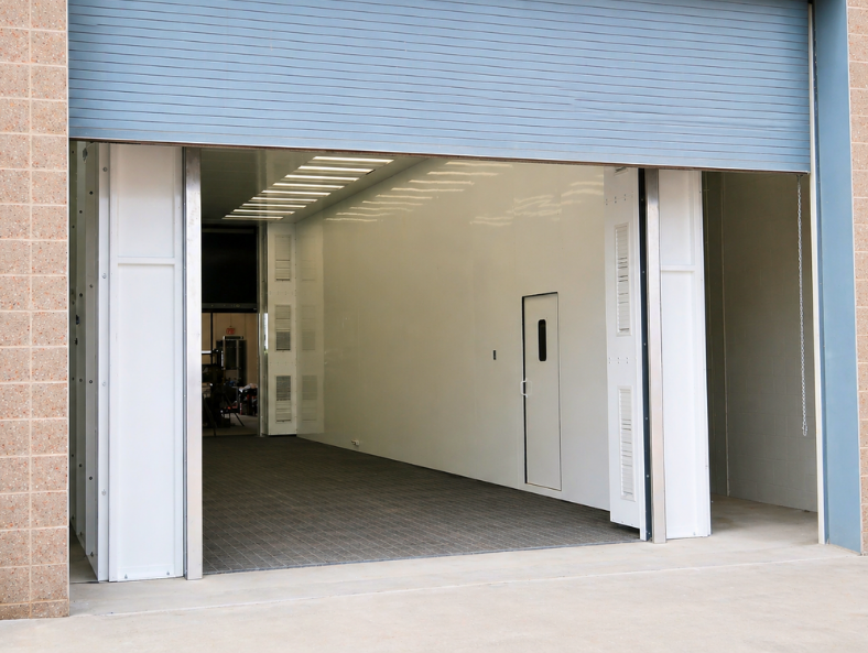 Large white industrial drive-thru blast enclosure with overhead door installed inside a facility.