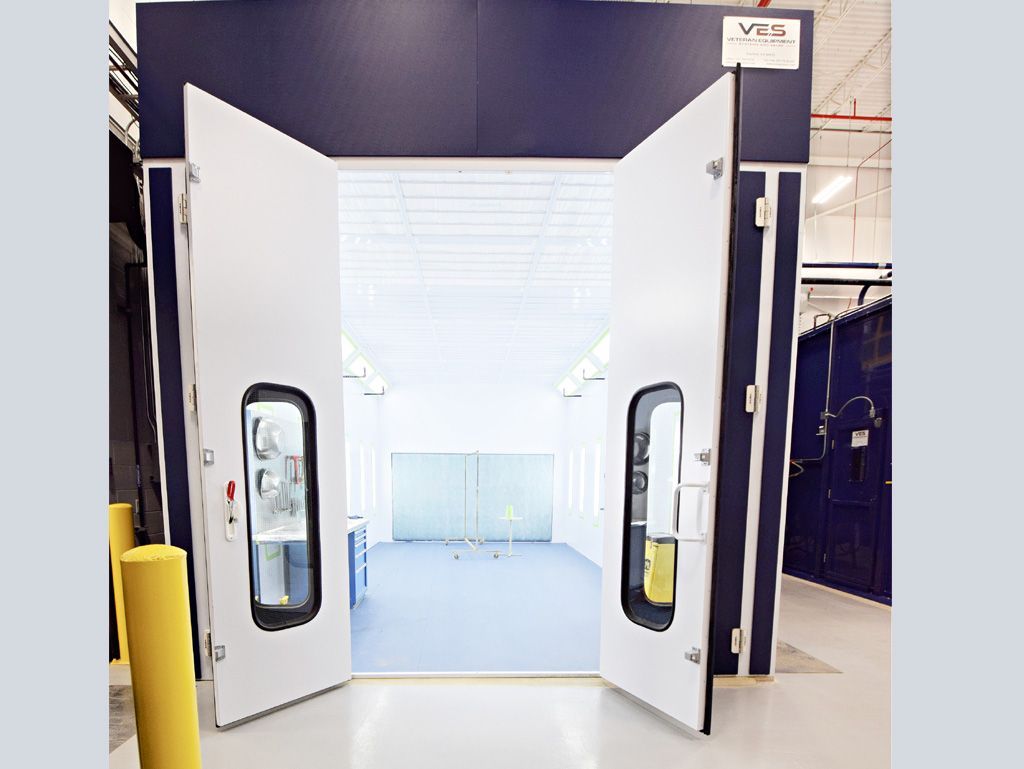 Spray booth with open white doors, blue accents, and a light interior. Industrial setting.