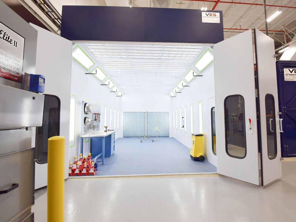 Open white paint booth with large doors, bright lights, and a blue floor, indoors.
