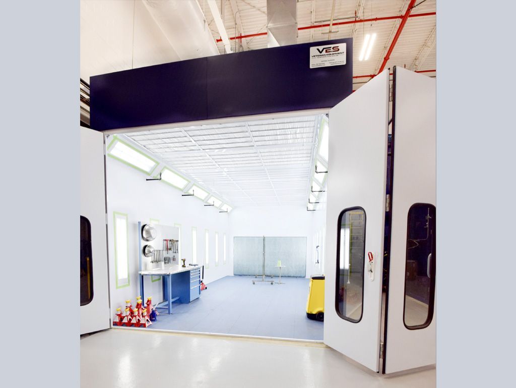 White and blue car paint booth in an industrial setting; open doorway, overhead lighting, blue flooring.