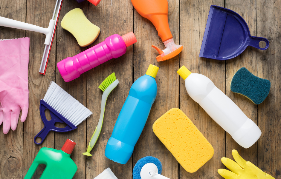 Een verzameling schoonmaakspullen, waaronder flessen, borstels, sponzen en handschoenen, uitgestald op een houten oppervlak.