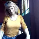 Persona con una blusa color amarillo mostaza sentada junto a una ventana con un fondo oscuro y una luz brillante.