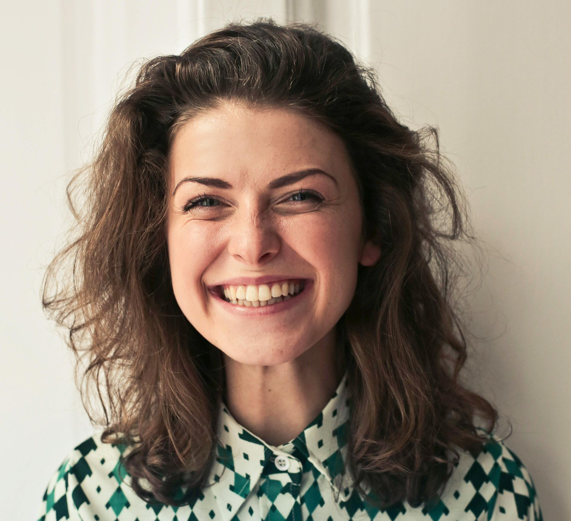 Woman with brown hair smiles broadly, wearing a patterned green and white shirt, against a white wall.