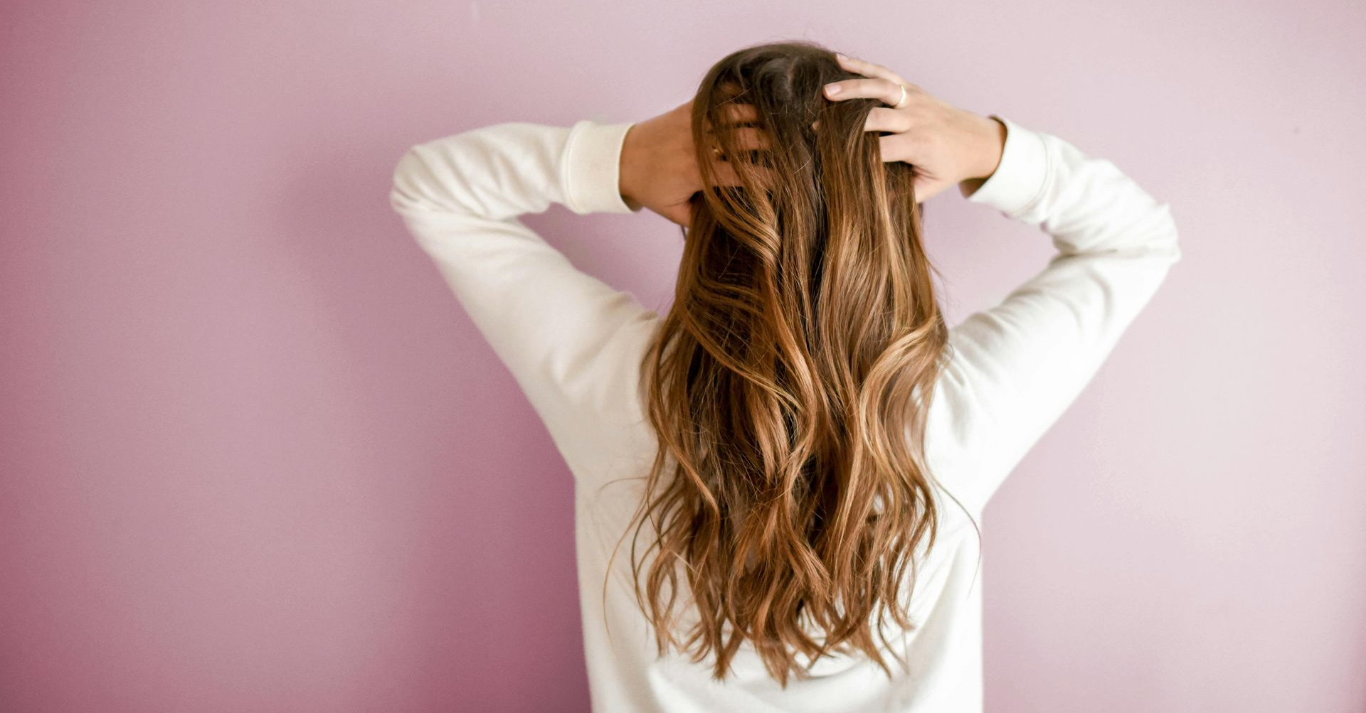 Woman with long, wavy brown hair, hands in hair, standing in front of a pink wall.